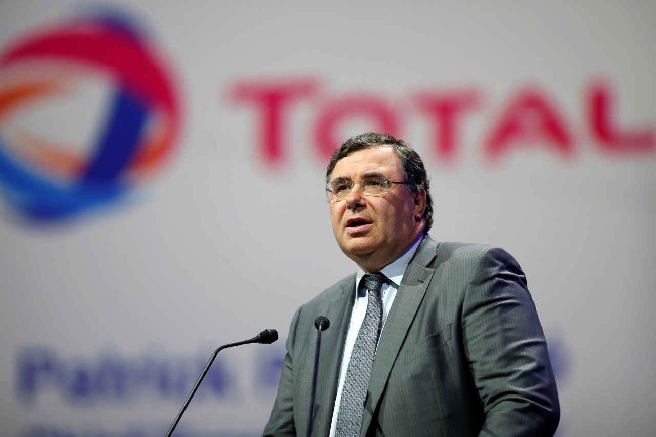 Man in a gray suit and blue patterned tie speaks at a podium during a conference, with a blurred red and blue logo in the background.