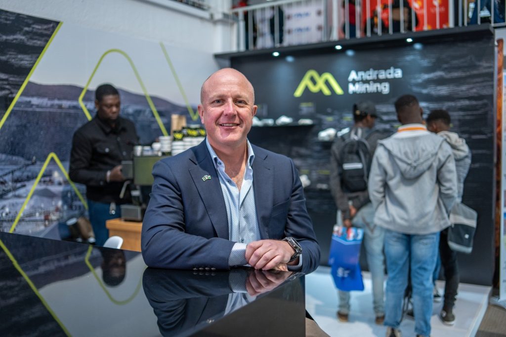 Anthony Viljoen | Mining and Energy Namibia | Namibia’s Leading Mining & Energy News Man in a navy suit standing at a glossy black booth counter, smiling at camera, Andrada Mining backdrop behind him.