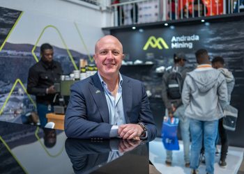 Anthony Viljoen | Mining and Energy Namibia | Namibia’s Leading Mining & Energy News Man in a navy suit standing at a glossy black booth counter, smiling at camera, Andrada Mining backdrop behind him.