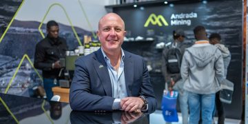Man in a navy suit standing at a glossy black booth counter, smiling at camera, Andrada Mining backdrop behind him.