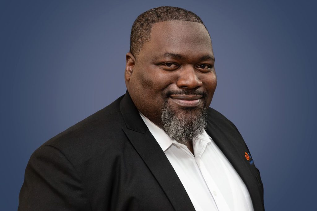 Professional headshot of a smiling Black man in a black suit jacket and white shirt against a blue gradient background.