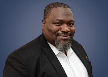 Professional headshot of a smiling Black man in a black suit jacket and white shirt against a blue gradient background.