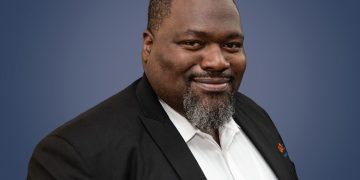 Irvinne Simataa | Mining and Energy Namibia | Namibia’s Leading Mining & Energy News Professional headshot of a smiling Black man in a black suit jacket and white shirt against a blue gradient background.