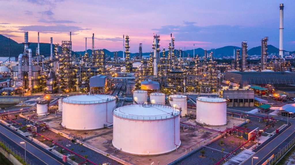 WhatsApp Image 2026-09-06 at 83353 PM | Mining and Energy Namibia | Namibia’s Leading Mining & Energy News Oil refinery at dusk: white storage tanks in foreground and a dense network of pipes and distillation columns under a purple sky with hills in the distance.