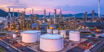 Oil refinery at dusk: white storage tanks in foreground and a dense network of pipes and distillation columns under a purple sky with hills in the distance.