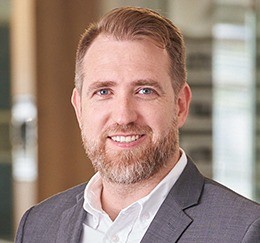 Portrait of a smiling man in a gray suit jacket and white shirt, office background, professional headshot.