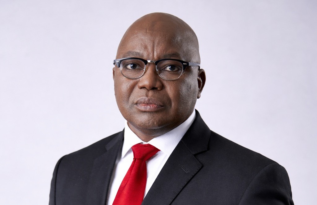 Professional head-and-shoulders portrait of a bald Black man in a black suit and red tie, wearing glasses.