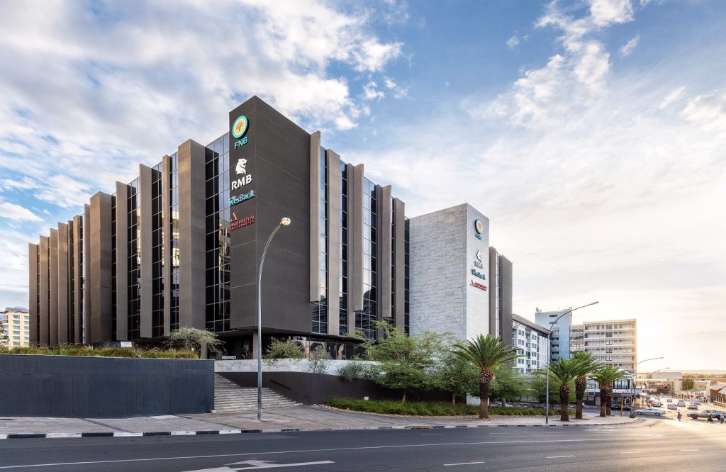 FNB BUILDING | Mining and Energy Namibia | Namibia’s Leading Mining & Energy News Modern office building with vertical concrete fins, logos along the facade, and palm trees lining the street.