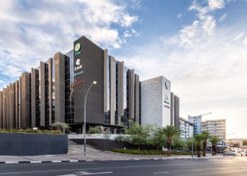 Modern office building with vertical concrete fins, logos along the facade, and palm trees lining the street.