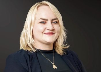 Stefanie Busch | Mining and Energy Namibia | Namibia’s Leading Mining & Energy News Smiling blonde woman in a dark blazer, wearing a pendant necklace, against a dark gradient background.