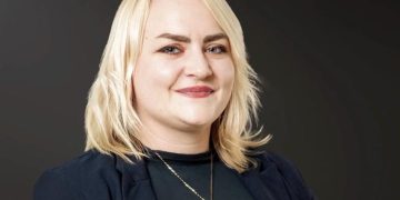 Stefanie Busch | Mining and Energy Namibia | Namibia’s Leading Mining & Energy News Smiling blonde woman in a dark blazer, wearing a pendant necklace, against a dark gradient background.