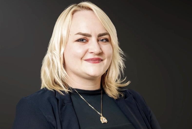 Smiling blonde woman in a dark blazer, wearing a pendant necklace, against a dark gradient background.