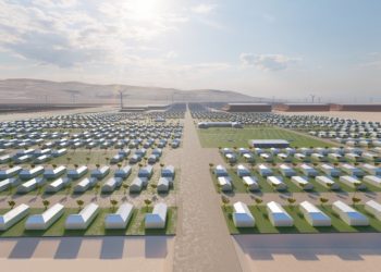 Desert development with hundreds of white rectangular buildings in a grid, a central road, and wind turbines on the horizon.