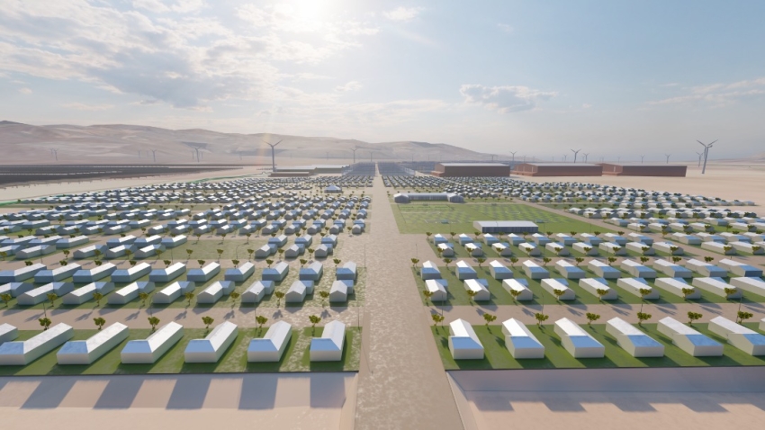 Desert development with hundreds of white rectangular buildings in a grid, a central road, and wind turbines on the horizon.