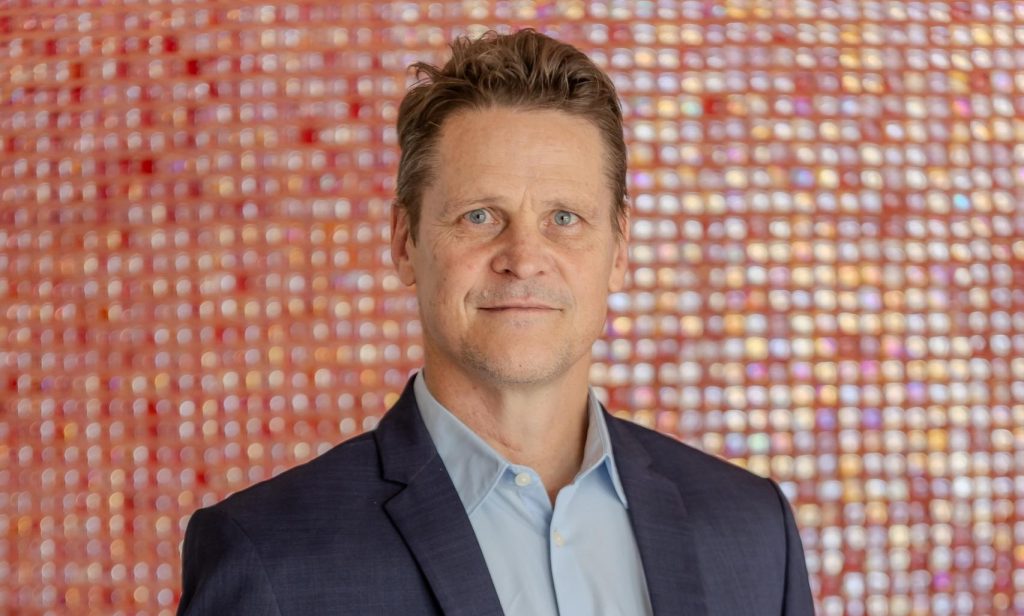 Riaan Vermeulen | Mining and Energy Namibia | Namibia’s Leading Mining & Energy News Portrait of a man in a dark suit and light blue shirt, standing against a colorful blurred mosaic background.