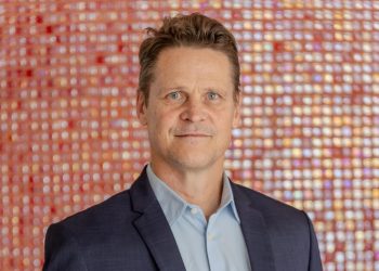 Riaan Vermeulen | Mining and Energy Namibia | Namibia’s Leading Mining & Energy News Portrait of a man in a dark suit and light blue shirt, standing against a colorful blurred mosaic background.