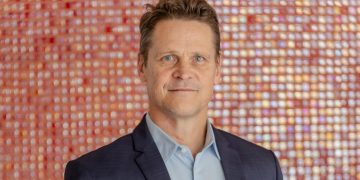 Riaan Vermeulen | Mining and Energy Namibia | Namibia’s Leading Mining & Energy News Portrait of a man in a dark suit and light blue shirt, standing against a colorful blurred mosaic background.