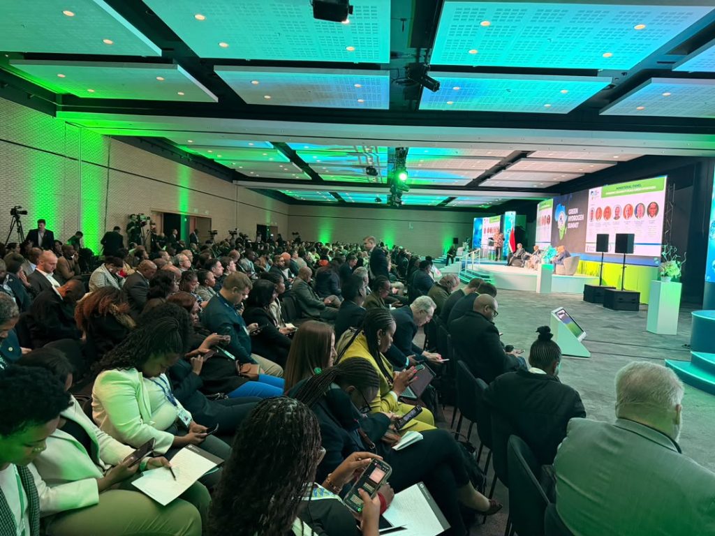 Large conference hall with green lighting, audience seated facing a panel on stage with microphones and screens in the background.