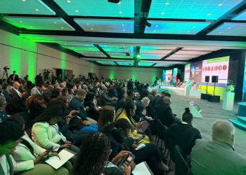 Large conference hall with green lighting, audience seated facing a panel on stage with microphones and screens in the background.