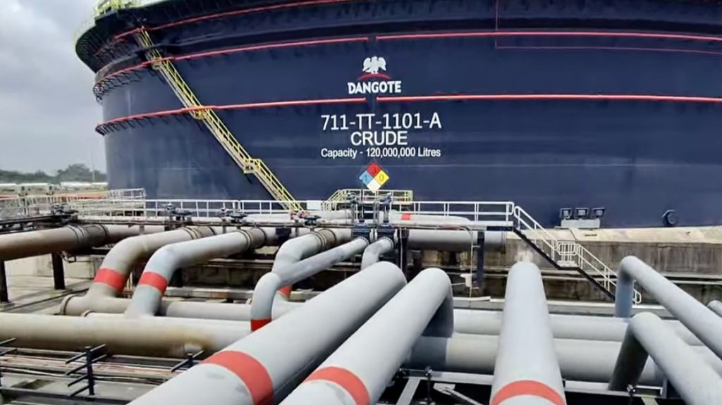 DANGOTE6 | Mining and Energy Namibia | Namibia’s Leading Mining & Energy News Massive blue crude oil storage tank labeled Dangote, with a yellow ladder and surrounding network of gray pipelines. A safety sign is visible in front of the tank far above a concrete platform.