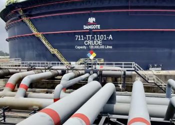 Massive blue crude oil storage tank labeled Dangote, with a yellow ladder and surrounding network of gray pipelines. A safety sign is visible in front of the tank far above a concrete platform.