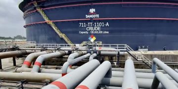 DANGOTE6 | Mining and Energy Namibia | Namibia’s Leading Mining & Energy News Massive blue crude oil storage tank labeled Dangote, with a yellow ladder and surrounding network of gray pipelines. A safety sign is visible in front of the tank far above a concrete platform.