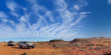 Two white off-road vehicles parked on red desert sand under a bright blue sky with wispy clouds, distant rock formations on the horizon.
