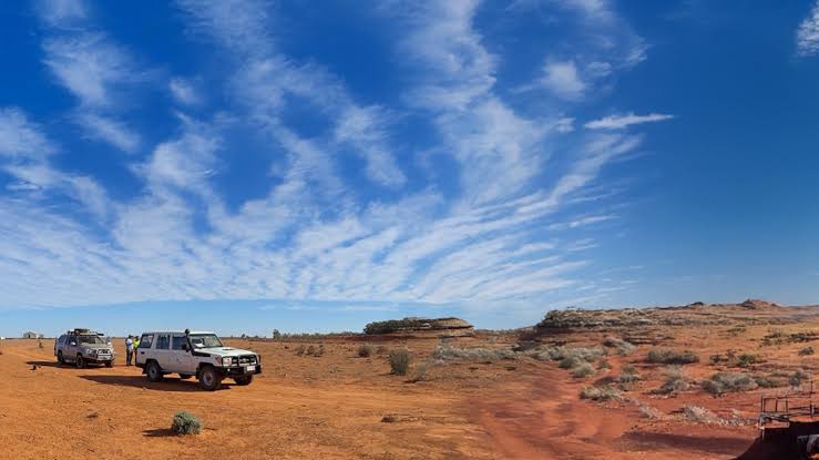 SULTAN RESOURCES | Mining and Energy Namibia | Namibia’s Leading Mining & Energy News Two white off-road vehicles parked on red desert sand under a bright blue sky with wispy clouds, distant rock formations on the horizon.