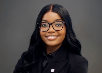 Professional headshot of a smiling woman with long black hair and glasses, wearing a black blazer against a gray backdrop.