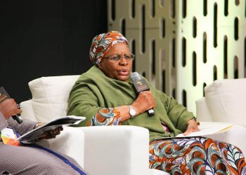 Panelist in a green shawl and colorful headwrap speaks into a handheld microphone, holding papers.