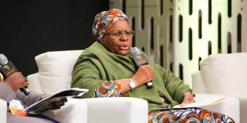 Panelist in a green shawl and colorful headwrap speaks into a handheld microphone, holding papers.