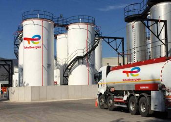 Industrial facility with tall white TotalEnergies storage tanks and a branded tanker truck in the foreground under a blue sky with stairs and piping visible.