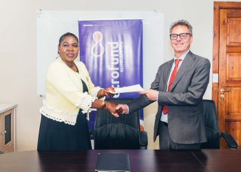 Two professionals shake hands across a dark conference table, exchanging a document in front of a blue banner.