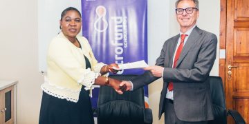Two professionals shake hands across a dark conference table, exchanging a document in front of a blue banner.