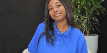 Young woman with long dark hair in a blue polo shirt, seated indoors with a plant behind her and a black wall backdrop.