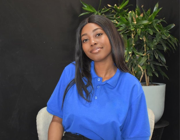 FRANSINA NELULU | Mining and Energy Namibia | Namibia’s Leading Mining & Energy News Young woman with long dark hair in a blue polo shirt, seated indoors with a plant behind her and a black wall backdrop.