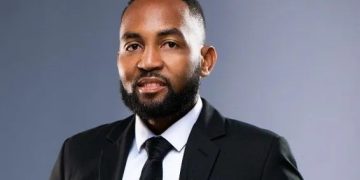 Professional headshot of a man in a black suit and tie, with a small flag lapel pin, against a gray gradient backdrop.