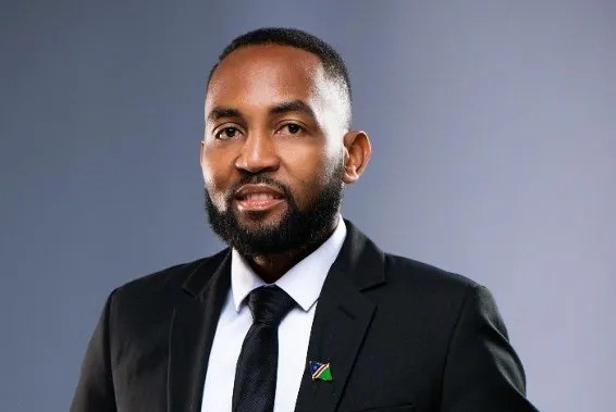 Professional headshot of a man in a black suit and tie, with a small flag lapel pin, against a gray gradient backdrop.