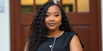Veripuami Tjipueja | Mining and Energy Namibia | Namibia’s Leading Mining & Energy News Portrait of a confident woman with long curly hair in a black sleeveless dress posed in front of a wooden door exterior.