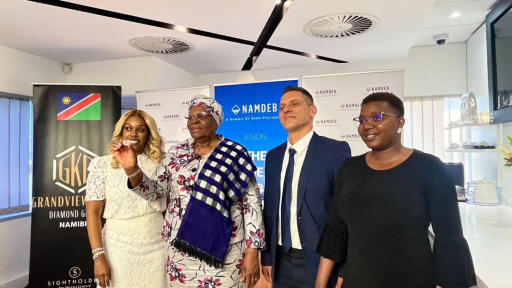 Four people stand in a corporate event space with Namdeb banners in the background; one woman in white holds up a small item for display.