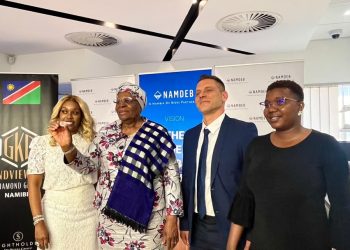 Four people stand in a corporate event space with Namdeb banners in the background; one woman in white holds up a small item for display.