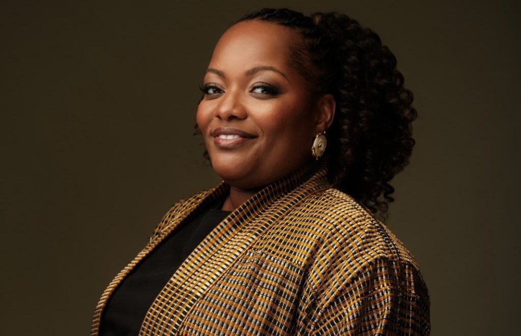 Portrait of a smiling Black woman with curly hair, wearing a tan plaid blazer and gold earrings against a dark backdrop.