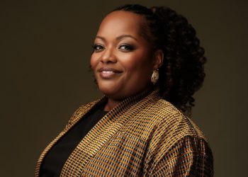 Portrait of a smiling Black woman with curly hair, wearing a tan plaid blazer and gold earrings against a dark backdrop.