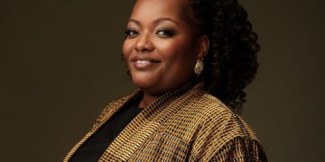 Portrait of a smiling Black woman with curly hair, wearing a tan plaid blazer and gold earrings against a dark backdrop.