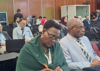 Front-row conference attendees: a woman in a green shawl writing with an orange pen, beside a man in a light gray suit and glasses.
