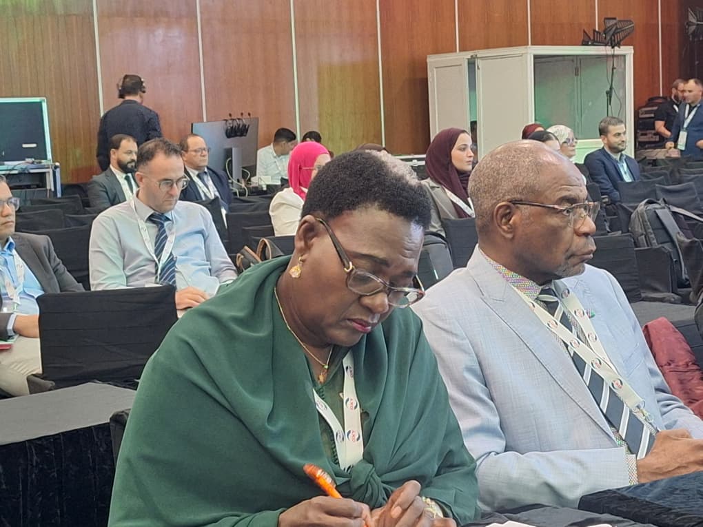 Front-row conference attendees: a woman in a green shawl writing with an orange pen, beside a man in a light gray suit and glasses.