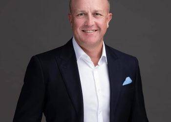Professional headshot: smiling man in a dark suit, white shirt, and light blue pocket square against a gray backdrop.