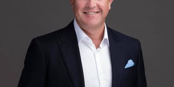 Professional headshot: smiling man in a dark suit, white shirt, and light blue pocket square against a gray backdrop.