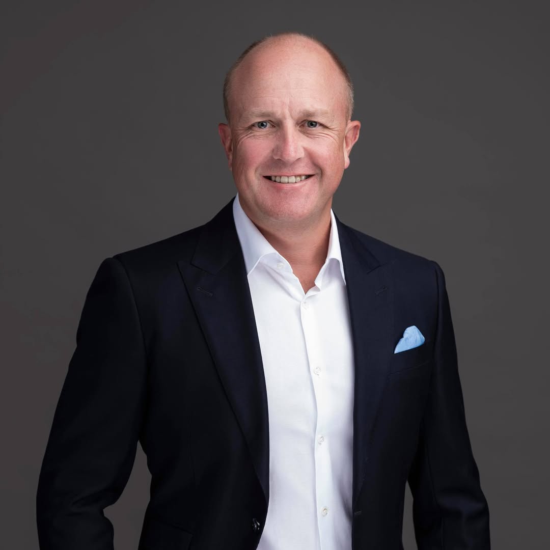 Professional headshot: smiling man in a dark suit, white shirt, and light blue pocket square against a gray backdrop.