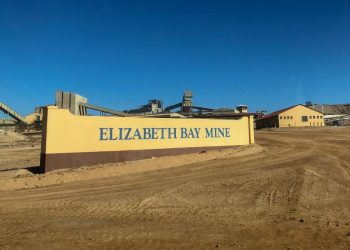 Entrance sign for Elizabeth Bay Mine with yellow wall and blue lettering, industrial buildings in the background under a clear blue sky.